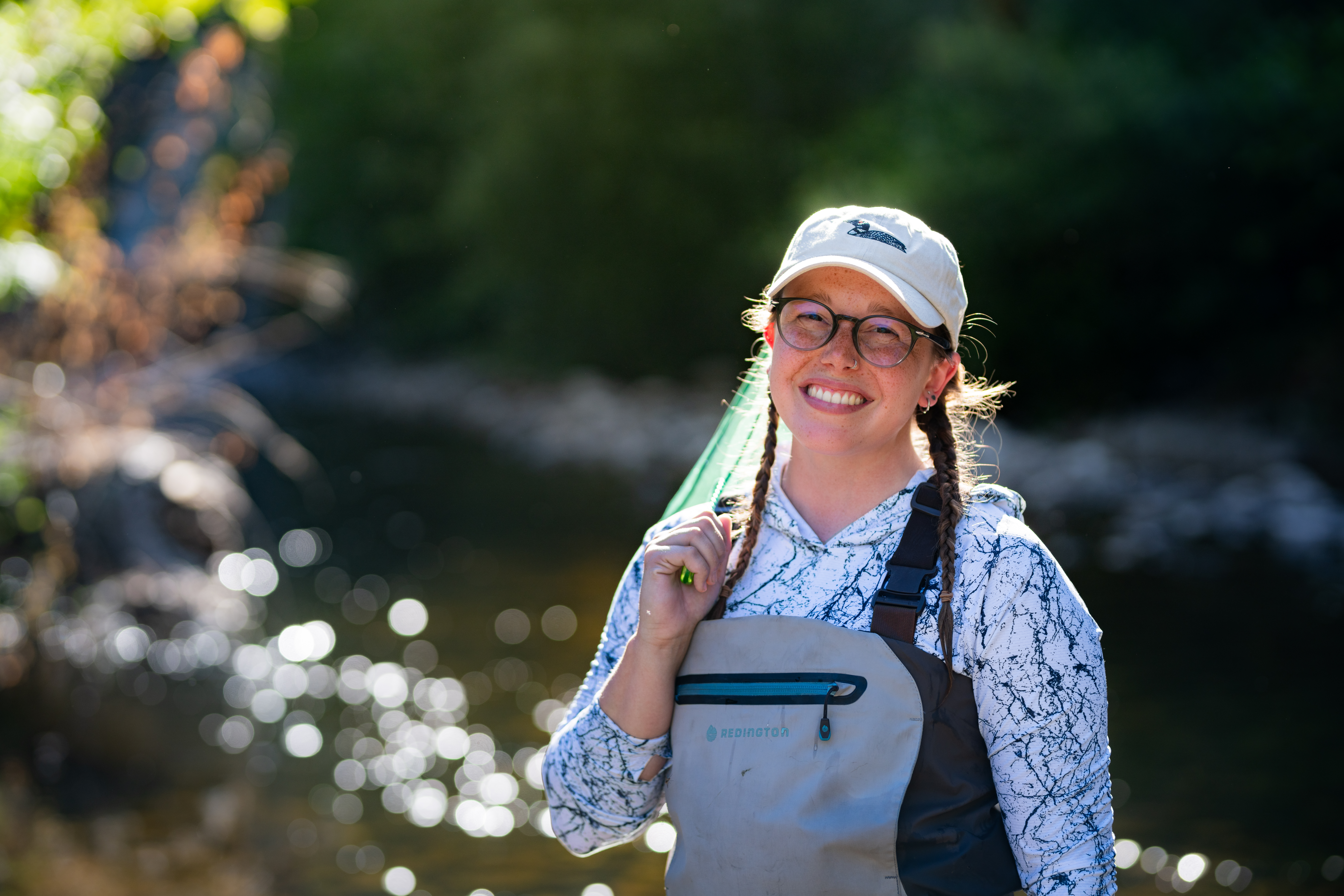Emily Correia-Lambton standing on a rock in a shallow stream wearing chest waders and holding a yellow field notebook while conducting outdoor environmental fieldwork in a wooded area